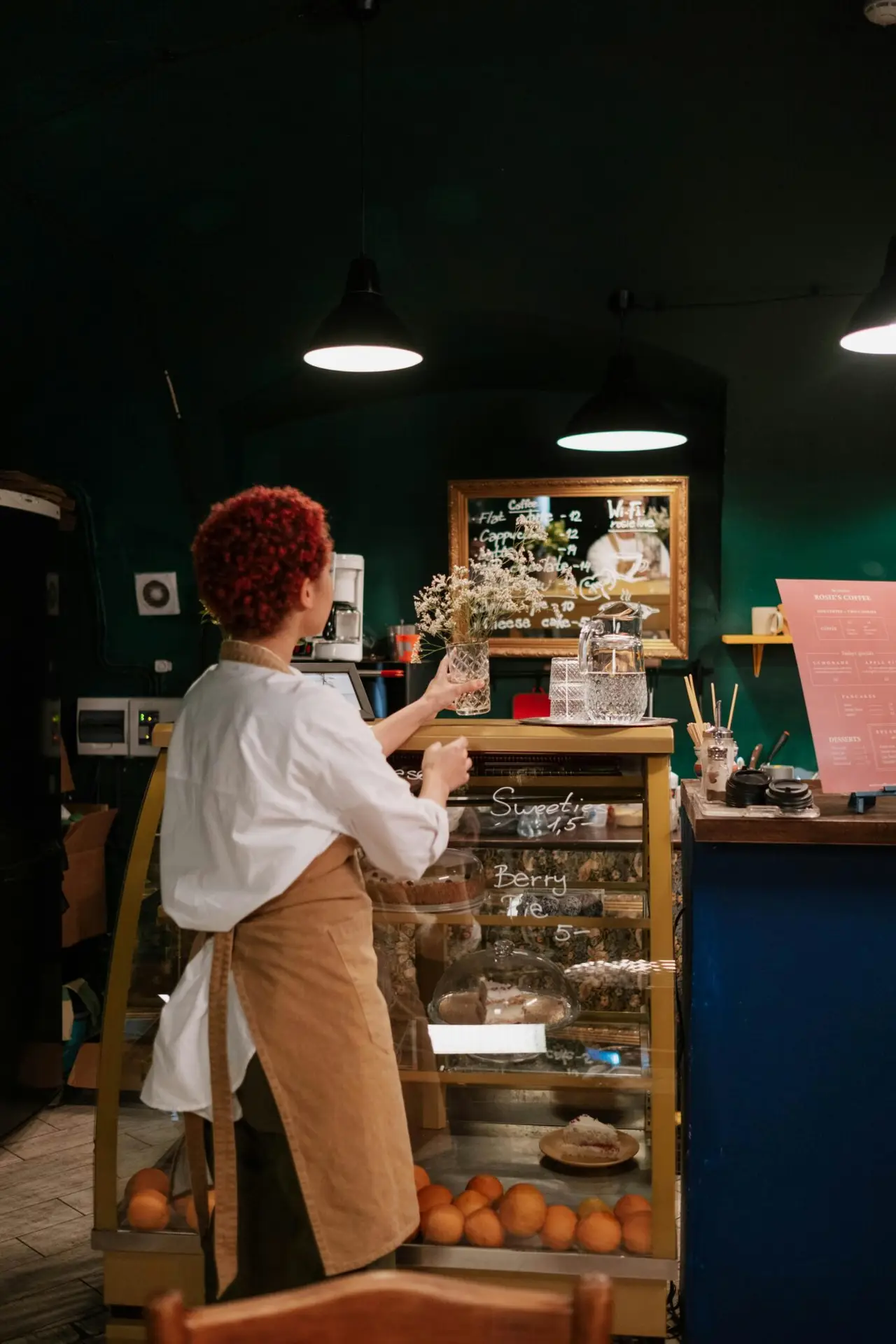 A waitress with short hair organizes a display in a cozy café. Indoor warm lighting.
